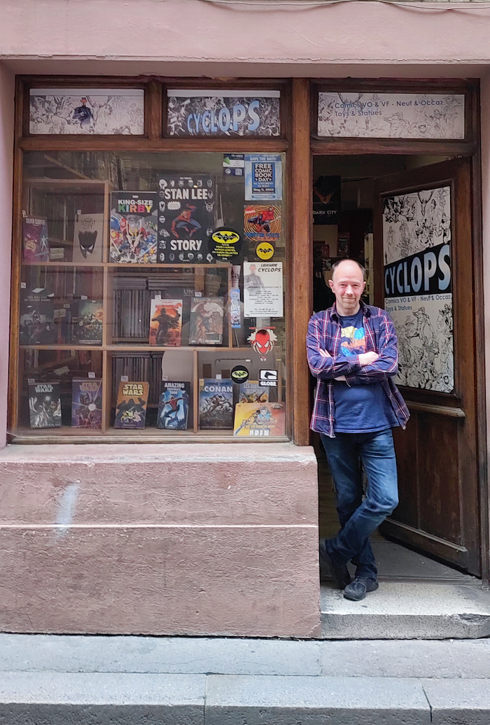 Photo d'un homme blanc en jeans et chemise à carreaux, accoudé à une devanture de magasin de comic-book. Derrière lui la porte est entrouverte et il est écrit Cyclops, c'est le nom du magasin.