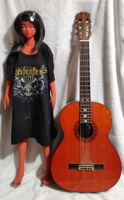 photo d'une poupée à la peau rouge et aux yeux dorés, elle porte un T-shirt noir beaucoup trop grand pour elle. Juste à coté d'elle à droite il y a une guitare classique qui est posée, elles font toutes les deux la même hauteur.